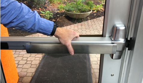 Image depicting a person's hand pressing down on a panic bar door opener, a safety feature commonly found on emergency exit doors.